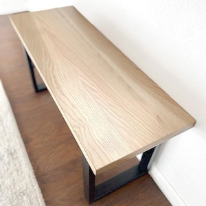 May include: A light-colored wooden bench with a rectangular top and black metal legs. The bench is positioned against a white wall and appears to be made of solid wood. The bench is approximately 48 inches long.