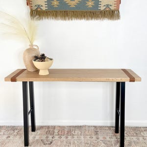 May include: A wooden console table with black metal legs and brown leather straps. The table is sitting on a rug with a geometric pattern.