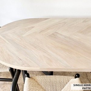 May include: Light wood oval dining table with a single herringbone pattern. The table is surrounded by two black chairs with woven seats. The text "SINGLE HERRINGBONE PATTERN" is visible.