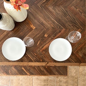 May include: Two white ceramic plates and two wine glasses sit on a dark brown wooden table with a herringbone pattern. Two white ceramic vases with a textured pattern are in the upper left corner of the image.