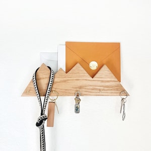 May include: A wooden key holder shaped like a mountain range with three hooks. There are keys and a lanyard hanging from the hooks. An envelope is placed in the top of the holder.