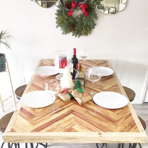 May include: A wooden dining table with a herringbone pattern top. The table is set with white plates, wine glasses, and a bottle of wine. There are also two small Christmas trees and wrapped gifts on the table.