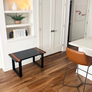 May include: A black rectangular bench with brown leather accents on the top edges. The bench has a minimalist design with a black metal frame. The room features a built-in white shelf with decor and a brown leather bar stool.