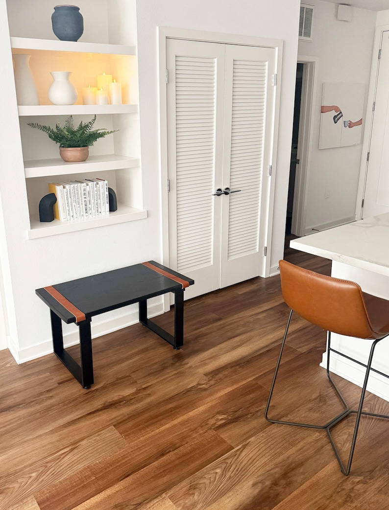 May include: A black rectangular bench with brown leather accents on the top edges. The bench has a minimalist design with a black metal frame. The room features a built-in white shelf with decor and a brown leather bar stool.