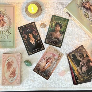 May include: An overhead shot of a collection of tarot cards, books, candles, crystals, and other mystical items. The central focus is on the "Mythos Tarot" deck, with several individual cards displayed. The color palette includes gold, cream, black, and various earth tones.