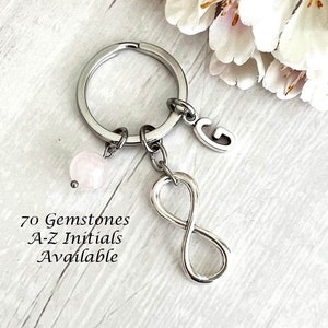 May include: A silver infinity charm keychain with a pink gemstone bead and a silver letter G charm. The text "70 Gemstones A-Z Initials Available" is printed below the keychain.