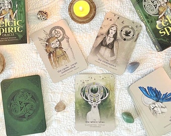 May include: A collection of spiritual items, including a "Celtic Spirit Oracle" card deck, candles, crystals, and other mystical objects. The items are arranged on a white quilted surface. The cards feature illustrations of mythical figures and are surrounded by other items.