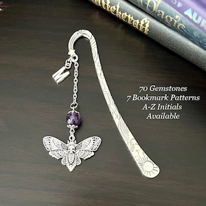 May include: A silver bookmark with a purple gemstone, a silver chain, and a silver moth charm. The bookmark is on a dark brown wooden surface. The text "70 Gemstones 7 Bookmark Patterns A-Z Initials Available" is visible in the image.