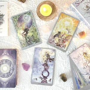 May include: An overhead shot of a collection of tarot cards, crystals, candles, and books. The cards are spread out on a white cloth, with the book titled "Shadowscapes Tarot" visible. Various crystals and candles surround the cards.
