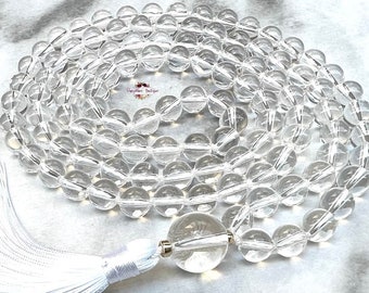May include: A mala necklace featuring numerous clear, spherical quartz beads arranged in a circular pattern. The necklace includes a larger, central clear bead and a long, white fabric tassel attached with a small metallic accent piece.