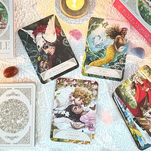 Op de afbeelding: Een verzameling tarotkaarten en een boek getiteld "Kate Forsyth's Fairytale Oracle" zijn gerangschikt op een witte doek. De kaarten bevatten illustraties van mythische figuren. Kaarsen, kristallen en andere mystieke items omringen de kaarten.