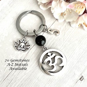 May include: A silver keychain with a black gemstone bead, a silver charm with a lotus flower and a meditating figure, and a silver charm with the Om symbol. The keychain also has a silver letter 'S' charm. The text '70 Gemstones A-Z Initials Available' is visible below the charms.