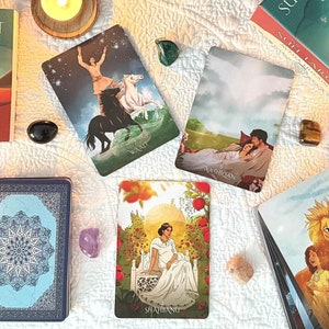 May include: A collection of tarot cards and a book titled "The Sufi Tarot" by Aveda Husain, arranged on a white cloth. The cards feature colorful illustrations, and lit candles and crystals surround the setup, creating a mystical atmosphere.