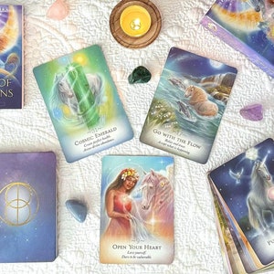 May include: A spread of oracle cards from the Magic of Unicorns deck, featuring images of unicorns, crystals, and celestial imagery. The cards are laid out on a white surface with candles and other magical objects.