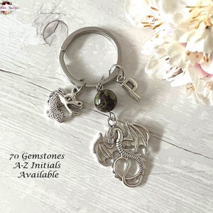 May include: A silver key chain with a dragon charm, a green gemstone bead, and a silver letter "P" charm. The text "70 Gemstones A-Z Initials Available" is printed on a wooden surface.