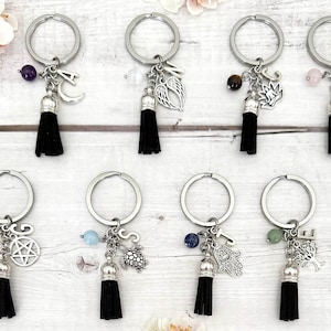 May include: A selection of silver keyrings, each adorned with a black tassel, gemstone, and a charm such as a moon or wings. The text states: "You will receive one key ring with your chosen gemstone, charm and initial."