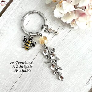May include: A silver keychain with a bee charm, a small flower charm, a clear gemstone, and a letter 'J' charm. The text '70 Gemstones A-Z Initials Available' is written below the keychain.