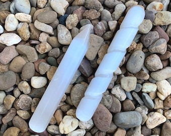 Puede incluir: Dos varitas de selenita blancas, una recta y otra retorcida, se muestran sobre un lecho de pequeñas piedras de colores. Las varitas son lisas y pulidas, con un color blanco lechoso natural.