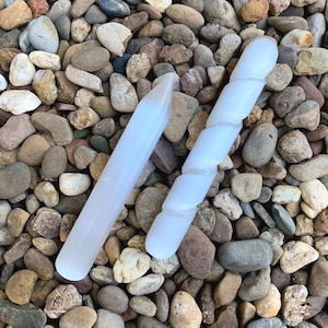 May include: Two white selenite wands, one straight and one twisted, are displayed on a bed of small, colourful stones. The wands are smooth and polished, with a natural, milky white colour.
