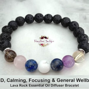 Puede incluir: Un brazalete difusor de aceites esenciales de roca de lava negra con cuentas de piedras preciosas marrones, rosas, azules, blancas y moradas. El brazalete está etiquetado como "TDAH, Calmante, Enfocando y Bienestar General".