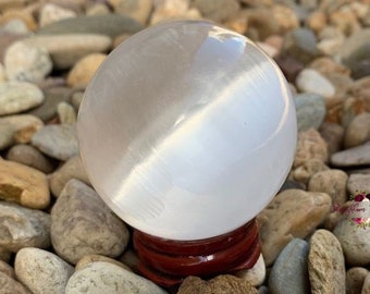 Puede incluir: Una esfera de cristal de selenita blanca, lisa y esférica, con un sutil brillo chatoyante, que descansa sobre un pequeño soporte de exhibición circular de madera de color marrón rojizo oscuro. La superficie está pulida y refleja la luz.