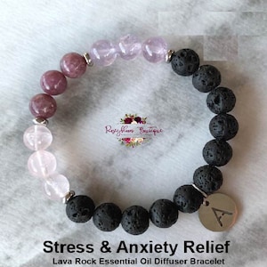 Pode incluir: Um bracelete difusor de óleo essencial de rocha de lava preta com contas de pedras preciosas rosa e roxas. O bracelete tem um amuleto de prata com a letra "A" nele. "Stress & Anxiety Relief" e "Lava Rock Essential Oil Diffuser Bracelet" estão escritos abaixo do bracelete.