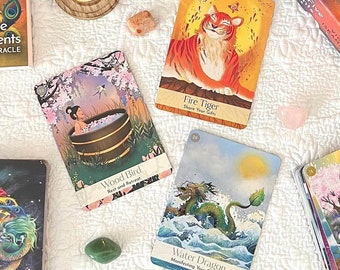 May include: A top-down view of a table with oracle cards, crystals, candles, and books. The cards display vibrant illustrations of a tiger, a dragon, and other elements. The book is titled "The Chinese Five Elements Oracle" by Vicki Iskandar.
