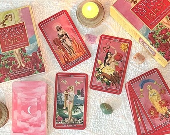 Puede incluir: Un conjunto de cartas del tarot y una caja, titulada "Goddess of Love Tarot", están dispuestas sobre una tela blanca. Las cartas presentan ilustraciones de mujeres y diseños florales. Velas, cristales y otros objetos místicos rodean las cartas del tarot.
