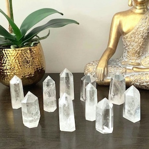 May include: A collection of clear quartz crystal points arranged on a wooden surface in front of a golden Buddha statue. The crystals are various sizes and shapes, with some being taller and thinner than others.