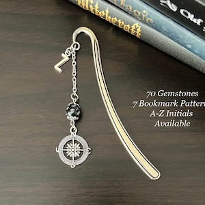 Puede incluir: Marcapáginas de plata con un dije de brújula y una cuenta de piedra preciosa negra. El marcapáginas tiene forma de un rectángulo largo y delgado con una parte superior curva. El texto "70 Gemstones", "7 Bookmark Patterns", "A-Z Initials" y "Available" está impreso en el marcapáginas.