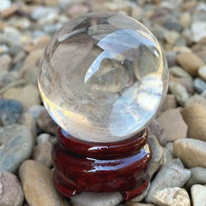 May include: A transparent crystal ball rests on a polished, dark red wooden stand. The sphere reflects the surroundings, and the stand has a rounded, tiered design. The background is a bed of small, smooth stones.