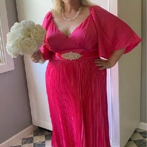 May include: A woman wearing a hot pink, pleated, long-sleeved dress with a matching sash and a rhinestone buckle. She is holding a bouquet of white flowers.