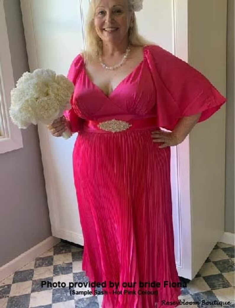 May include: A woman wearing a hot pink, pleated, long-sleeved dress with a matching sash and a rhinestone buckle. She is holding a bouquet of white flowers.