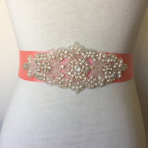 May include: A coral satin ribbon sash with a large, ornate embellishment featuring pearls and rhinestones.