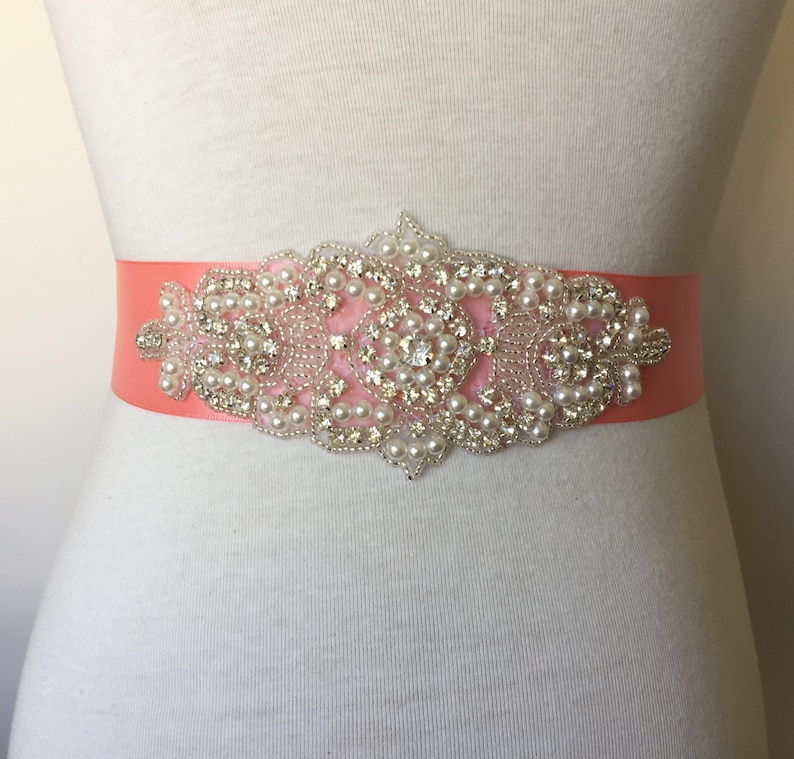 May include: A coral satin ribbon sash with a large, ornate embellishment featuring pearls and rhinestones.