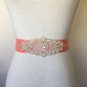 May include: A coral satin sash with a large, ornate rhinestone and pearl embellishment. The embellishment is silver and features a central round rhinestone surrounded by smaller rhinestones and pearls.