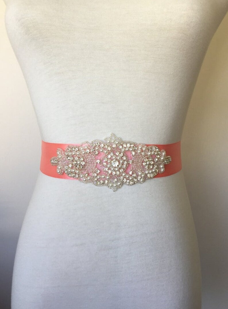 May include: A coral satin sash with a large, ornate rhinestone and pearl embellishment. The embellishment is silver and features a central round rhinestone surrounded by smaller rhinestones and pearls.