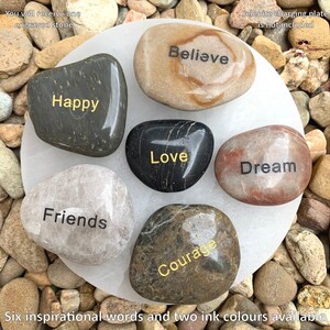 May include: Six smooth, round stones with inspirational words engraved in gold lettering: Believe, Happy, Love, Courage, Dream, and Friends. The stones are arranged on a white circular surface.