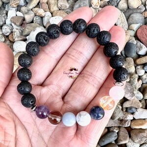 May include: A black lava rock bracelet with silver spacers and seven colorful gemstone beads. The bracelet is on a hand with a white background.