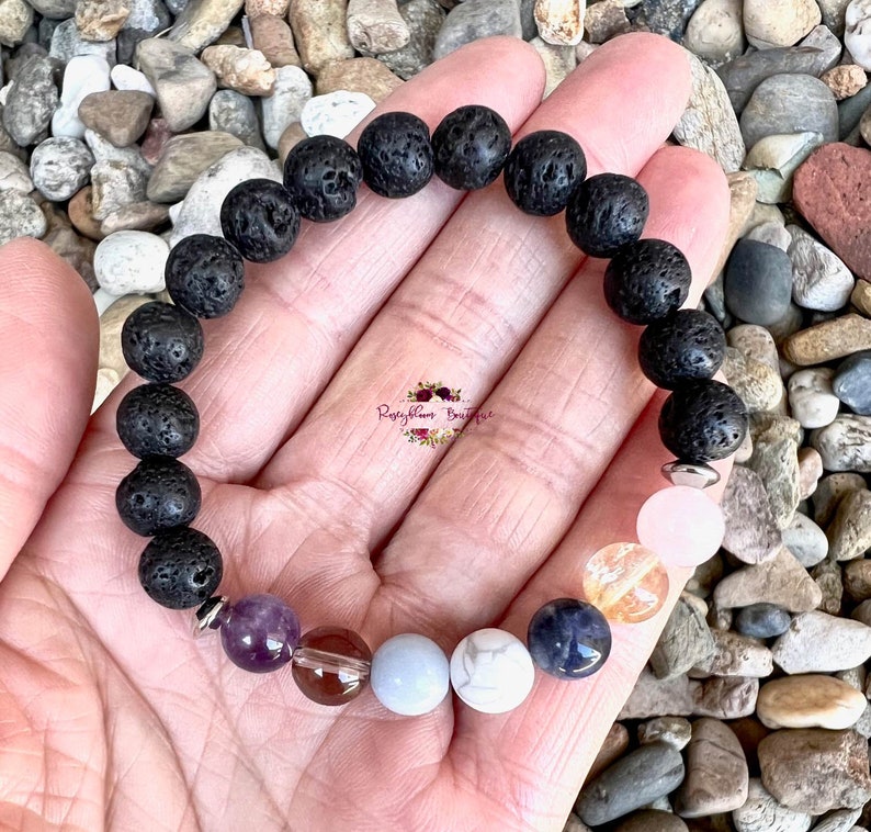 May include: A black lava rock bracelet with silver spacers and seven colorful gemstone beads. The bracelet is on a hand with a white background.