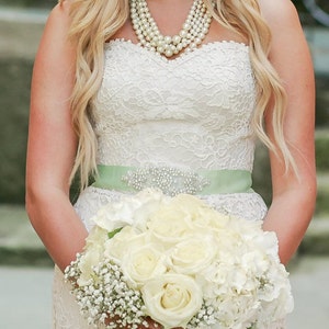 May include: A bride wearing a white lace wedding dress with a light green sash and a pearl necklace. She is holding a bouquet of white roses and baby's breath.