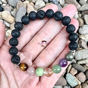 Puede incluir: Un brazalete de roca de lava negra con siete cuentas de piedras preciosas de colores. Las cuentas son verdes, naranjas, rosas, moradas y marrones. El brazalete est&aacute; en una mano.