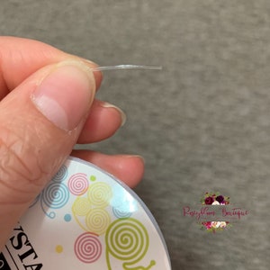 May include: A close-up of a clear, thin, flexible elastic cord held between two fingers. The cord is about 10 inches long.