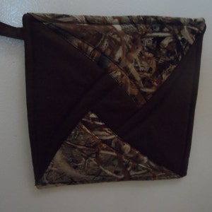 Pode incluir: Pega-panela quadrada marrom e com padrão de camuflagem com uma alça para pendurar. A pega-panela apresenta um tecido marrom escuro e um padrão de camuflagem em tons de marrom, bege e verde.