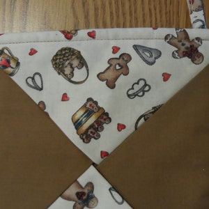 May include: A brown and white quilted pot holder with a gingerbread theme. The centre fabric features gingerbread men, hearts, and baking-related images. The edges are a solid brown colour.