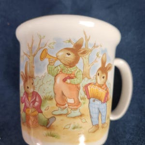 May include: A white ceramic mug featuring a colourful illustration of three anthropomorphic rabbits playing musical instruments in a wooded setting. One rabbit plays a trumpet, another a drum, and the third plays a yellow accordion.