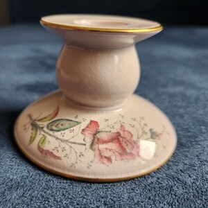May include: A cream-coloured ceramic candle holder with a subtle crackle glaze finish. The base features a delicate floral design with pink roses and green leaves. The top rim and the base edge are accented with a thin, metallic gold trim.