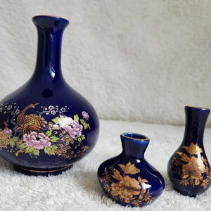 May include: Three cobalt blue ceramic vases with gold trim. The largest vase features a detailed peacock and floral design in gold, pink, and green. The two smaller vases are decorated with gold floral patterns. All three have narrow necks and rounded bodies.