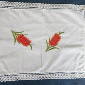 May include: A rectangular white table runner made of fabric featuring a delicate white lace border along the edges. The centre of the runner is decorated with two embroidered motifs of red bottlebrush flowers with green leaves and stems.