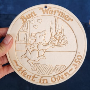 May include: A round, beige ceramic bun warmer featuring an embossed illustration of a pig holding a tray of buns. The text reads Bun Warmer at the top and Heat in Oven 350 degrees at the bottom. It includes a small hole at the top for hanging.
